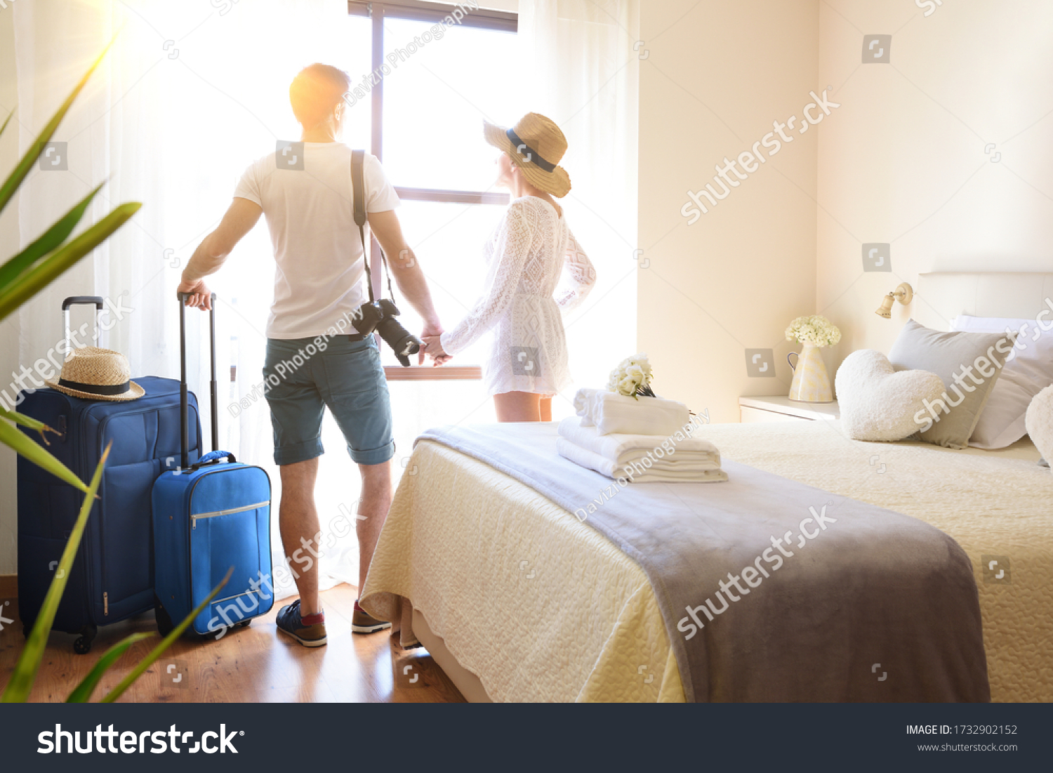 Couple on vacation with suitcases holding hands on their backs looking out a window in a bright hotel room. #1732902152