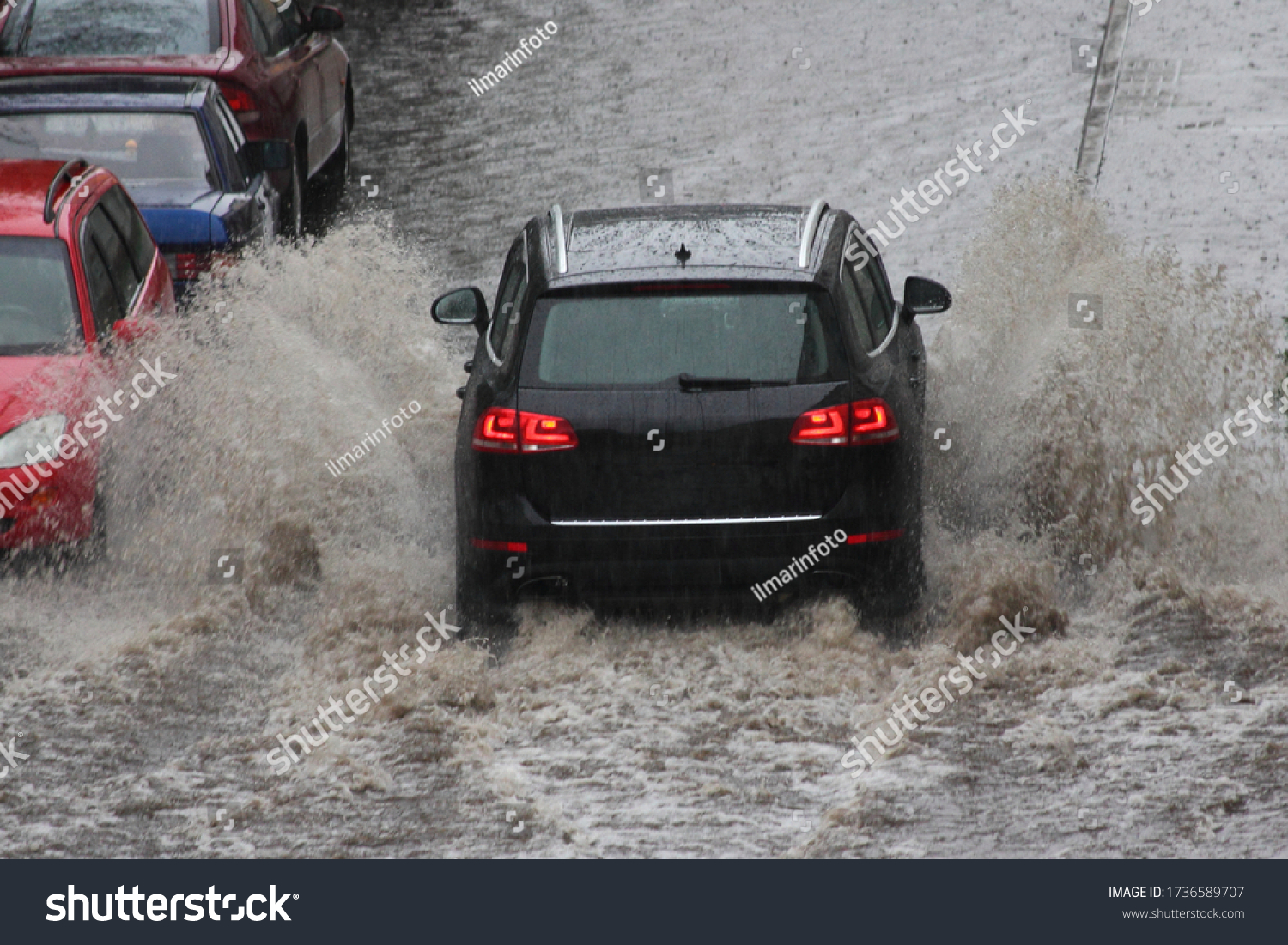 A black SUV car is driving fast with heavy spray in a deep puddle next to parked cars in heavy rain in the city on a spring day, the danger of hydraulic shock of engine, back view close up #1736589707