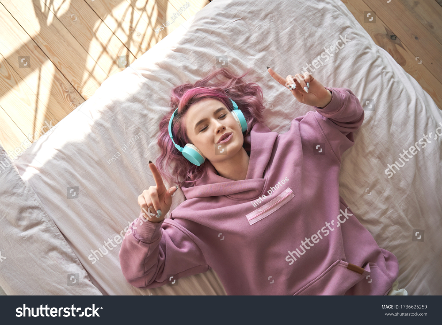 Happy funny teen girl with pink hair wear headphones lying in comfortable bed listening new pop music enjoying singing song with eyes closed relaxing in cozy bedroom at home. Top view from above. #1736626259