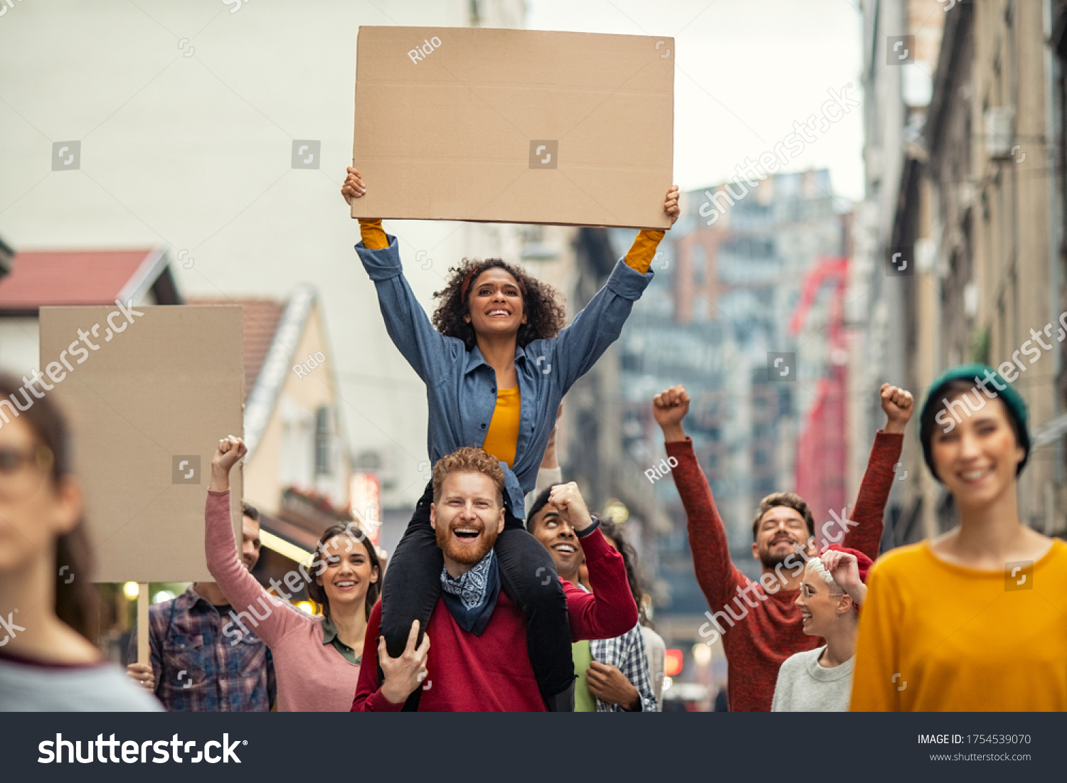 Group of multiethnic people on street holding blank cardboard placard celebrating victory during a protest. Group of content men and smiling women marching through a city. People protesting on road. #1754539070