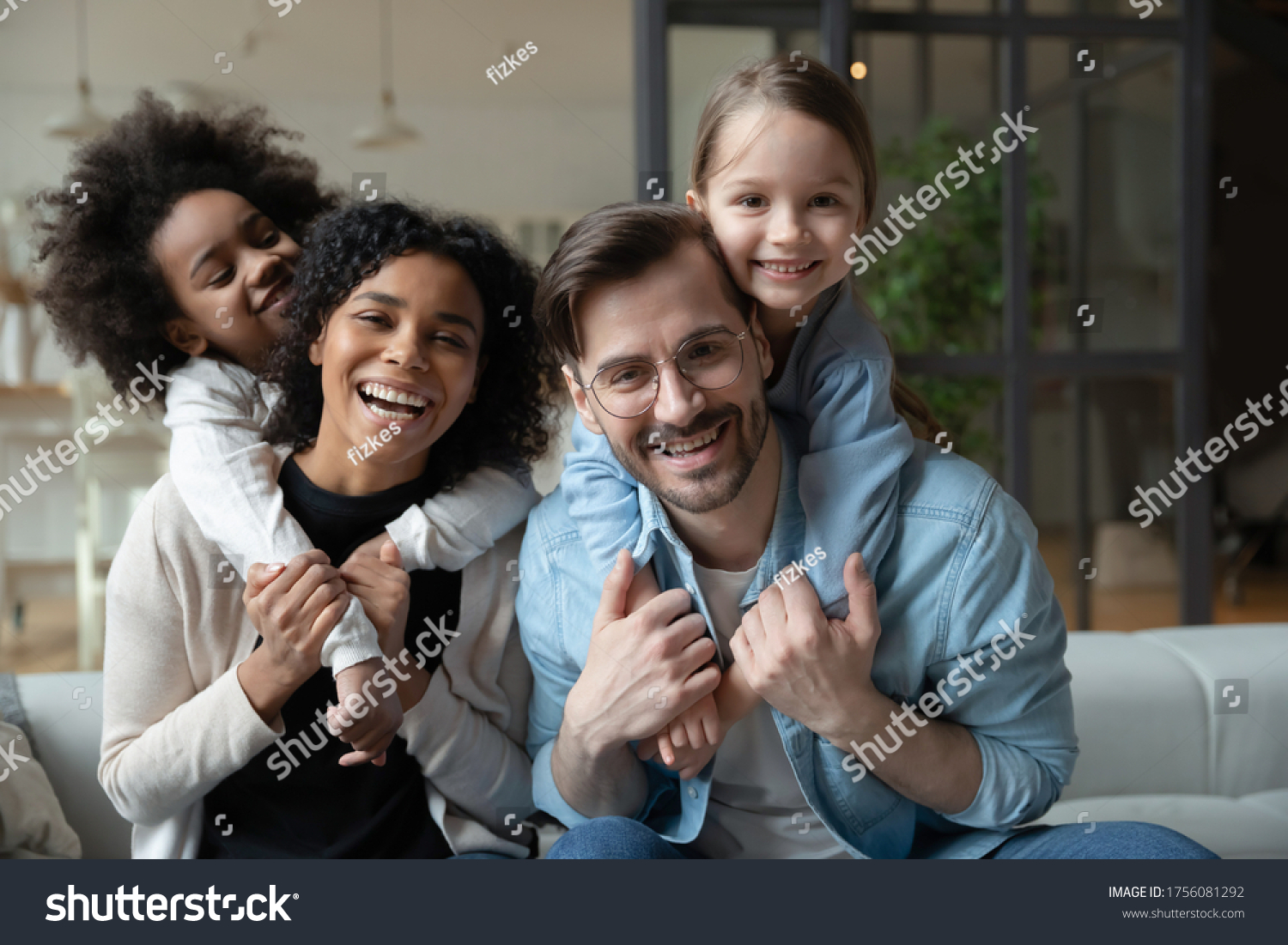 Cheery African and Caucasian girls cuddle piggy back multi-racial parents loving mom and dad seated on sofa in living room. Happy multinational family portrait, new home funny weekend together concept #1756081292