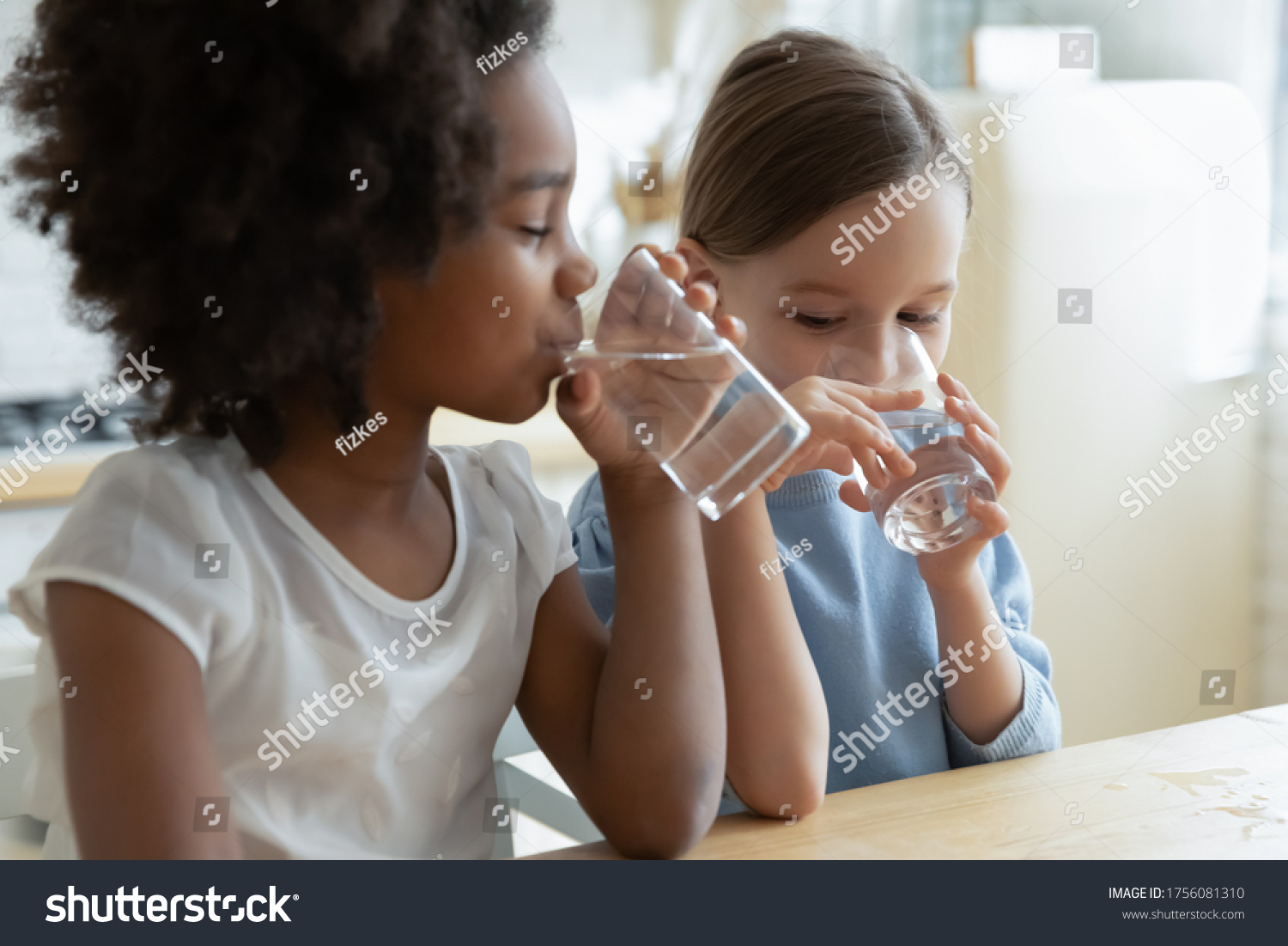 Two multi racial little girls sit at table in kitchen feels thirsty drink clean still natural or mineral water close up image. Healthy life habit of kids, health benefit dehydration prevention concept #1756081310