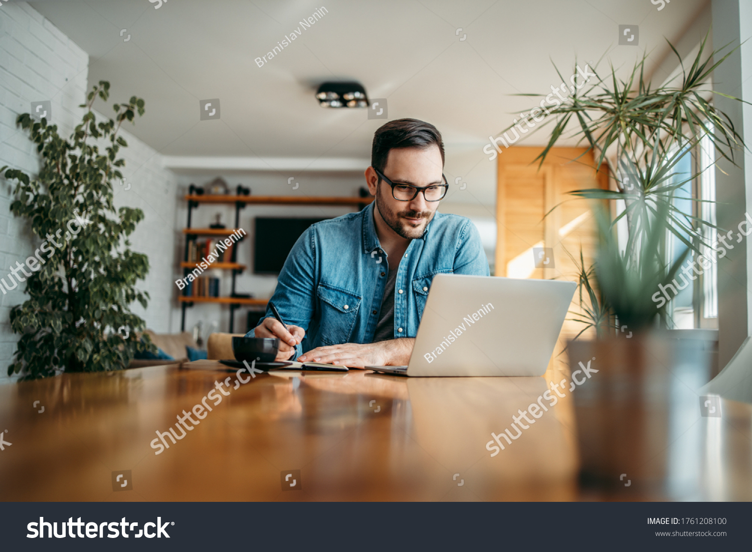 Handsome man taking notes and looking at laptop, at home office, portrait. #1761208100