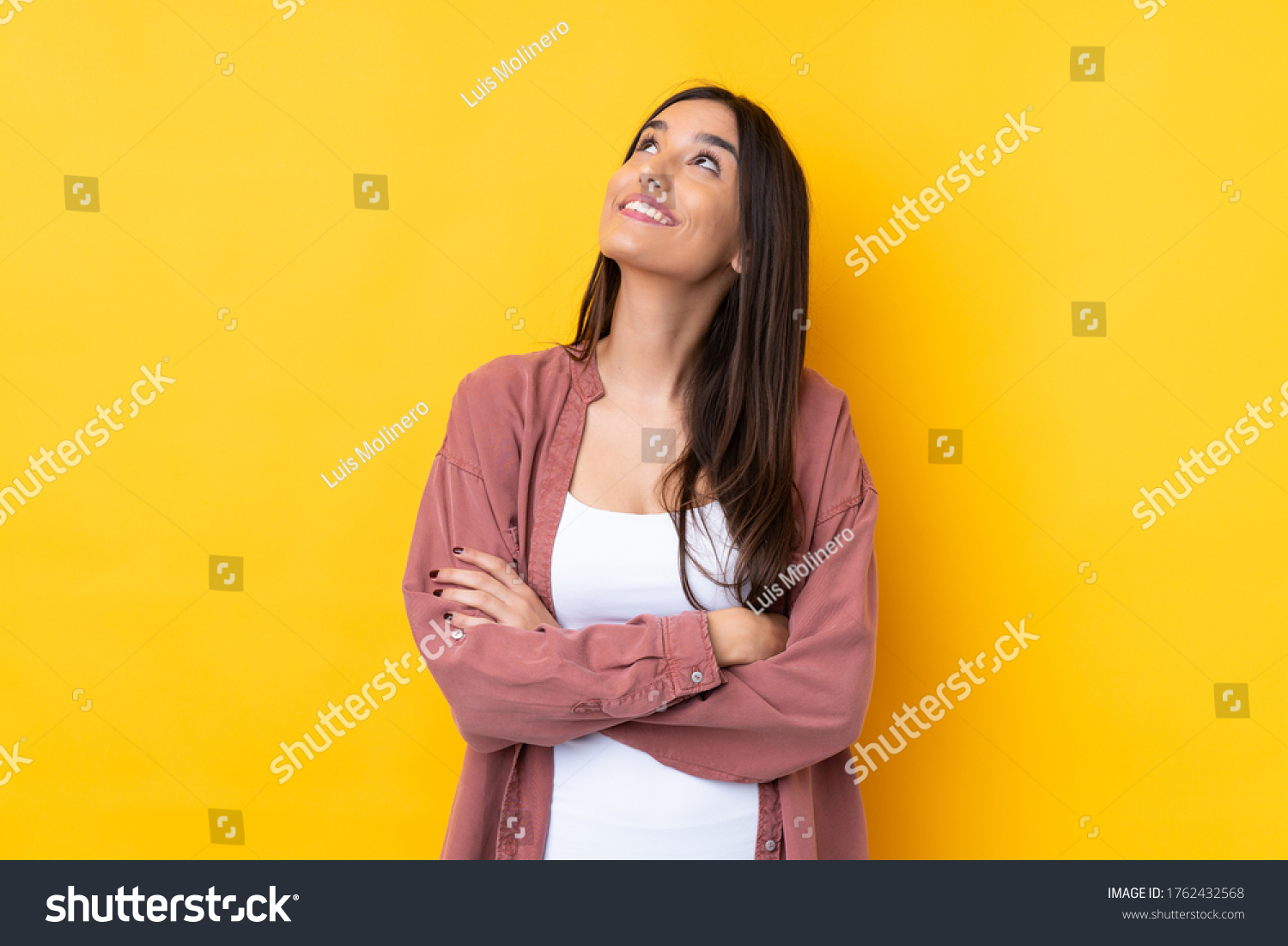 Young brunette woman over isolated yellow background looking up while smiling #1762432568
