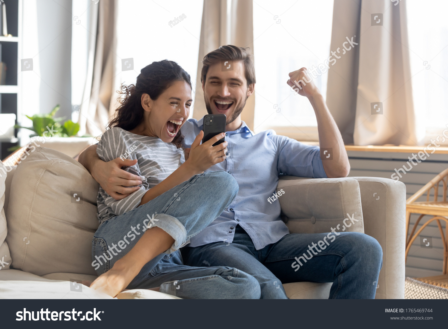 Overjoyed surprised young couple looking at phone screen, reading message with good news, excited woman and man celebrating success, online lottery win, showing yes gesture, sitting on couch #1765469744