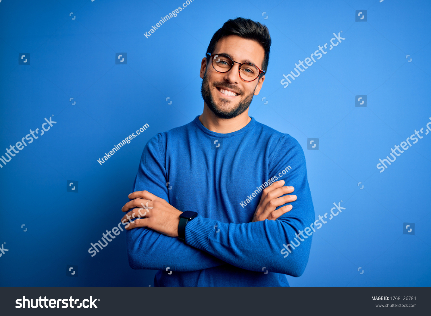 Young handsome man with beard wearing casual sweater and glasses over blue background happy face smiling with crossed arms looking at the camera. Positive person. #1768126784