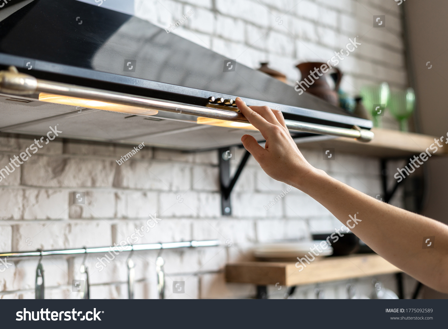 Cropped view of woman hand select mode on cooking hood, standing near kitchen appliance in contemporary interior with brick wall and decor on shelves at blurred background #1775092589