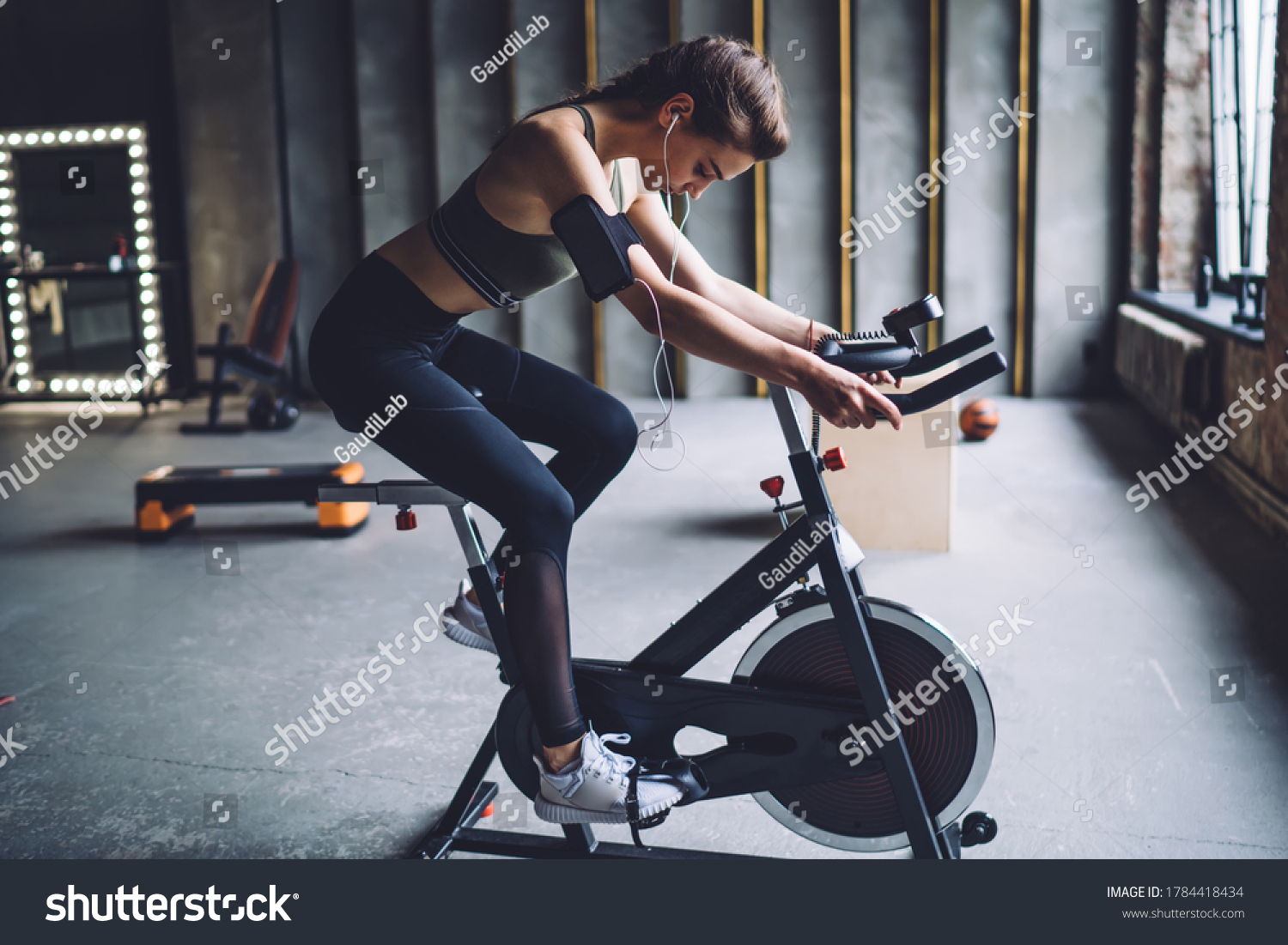 Concentrated fit female in sportswear with dark braided hair burning calories on spin bike and listening to music in headphones #1784418434