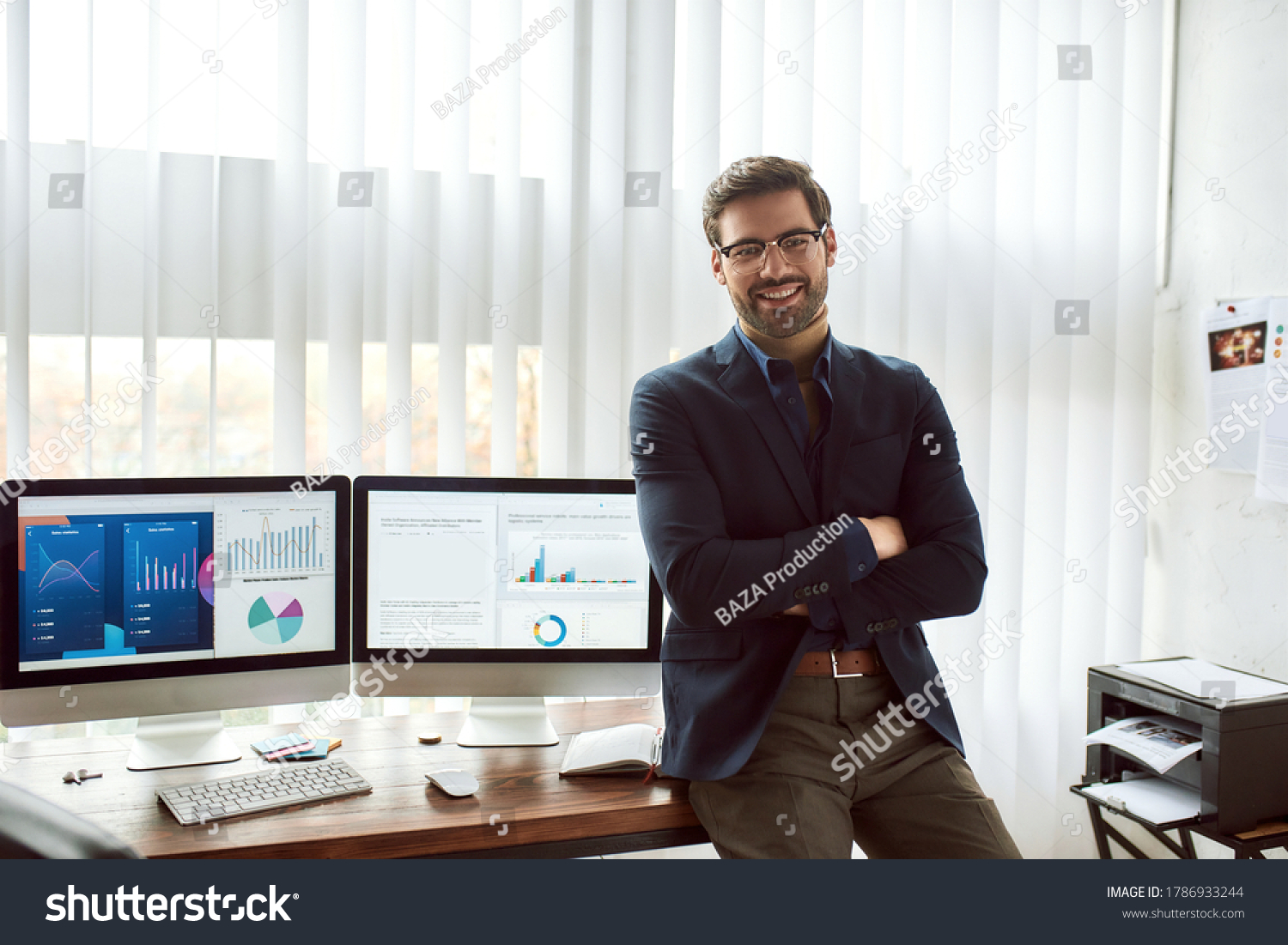 Young happy financial analyst in classic wear and eyeglasses leaning against a table and smiling at camera while working with statistical data, graphs and charts on computer in the modern office #1786933244