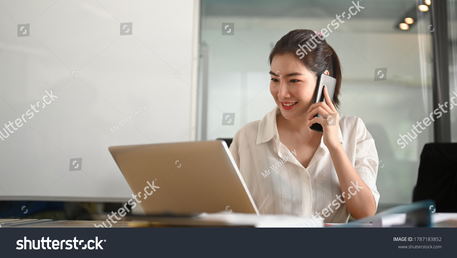 A businesswoman is talking on the mobile phone while sitting and using a computer laptop at the working desk. #1787183852