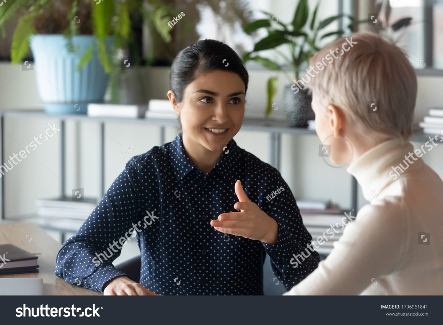 Friendly millennial indian businesswoman talking to blonde female colleague, sitting at table in office. Two young multiracial employees discussing working issues or enjoying informal conversation. #1796961841