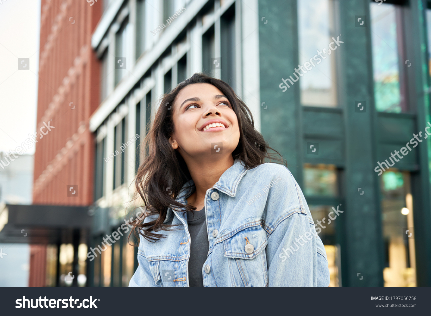 Confident happy beautiful young hipster African American woman wearing denim jacket looking up standing on city street outdoors dreaming, thinking or good future on urban buildings background. #1797056758