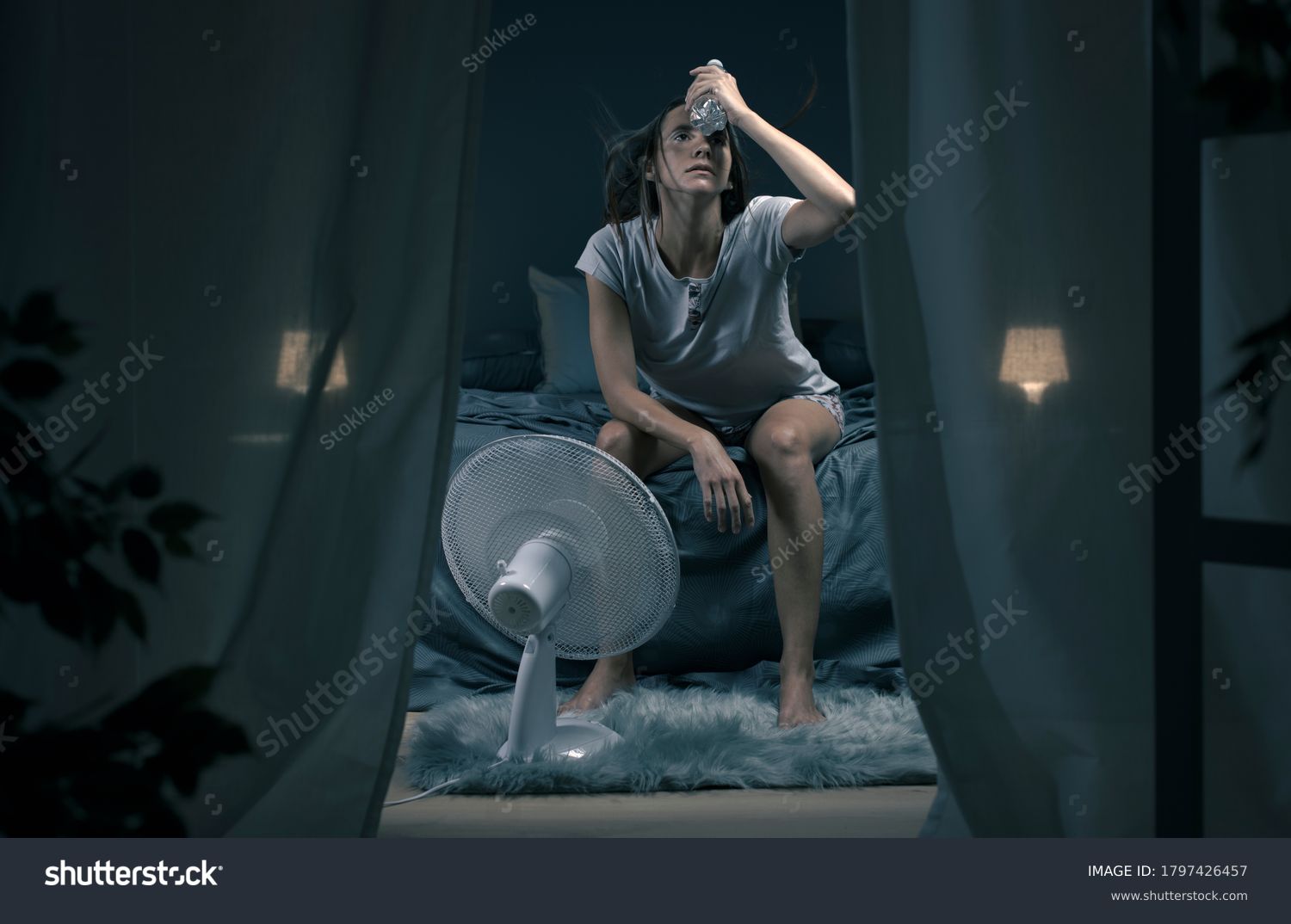 Exhausted woman suffering suring the heatwave, she is holding a water bottle and sitting in front of a cooling fan in the bedroom #1797426457