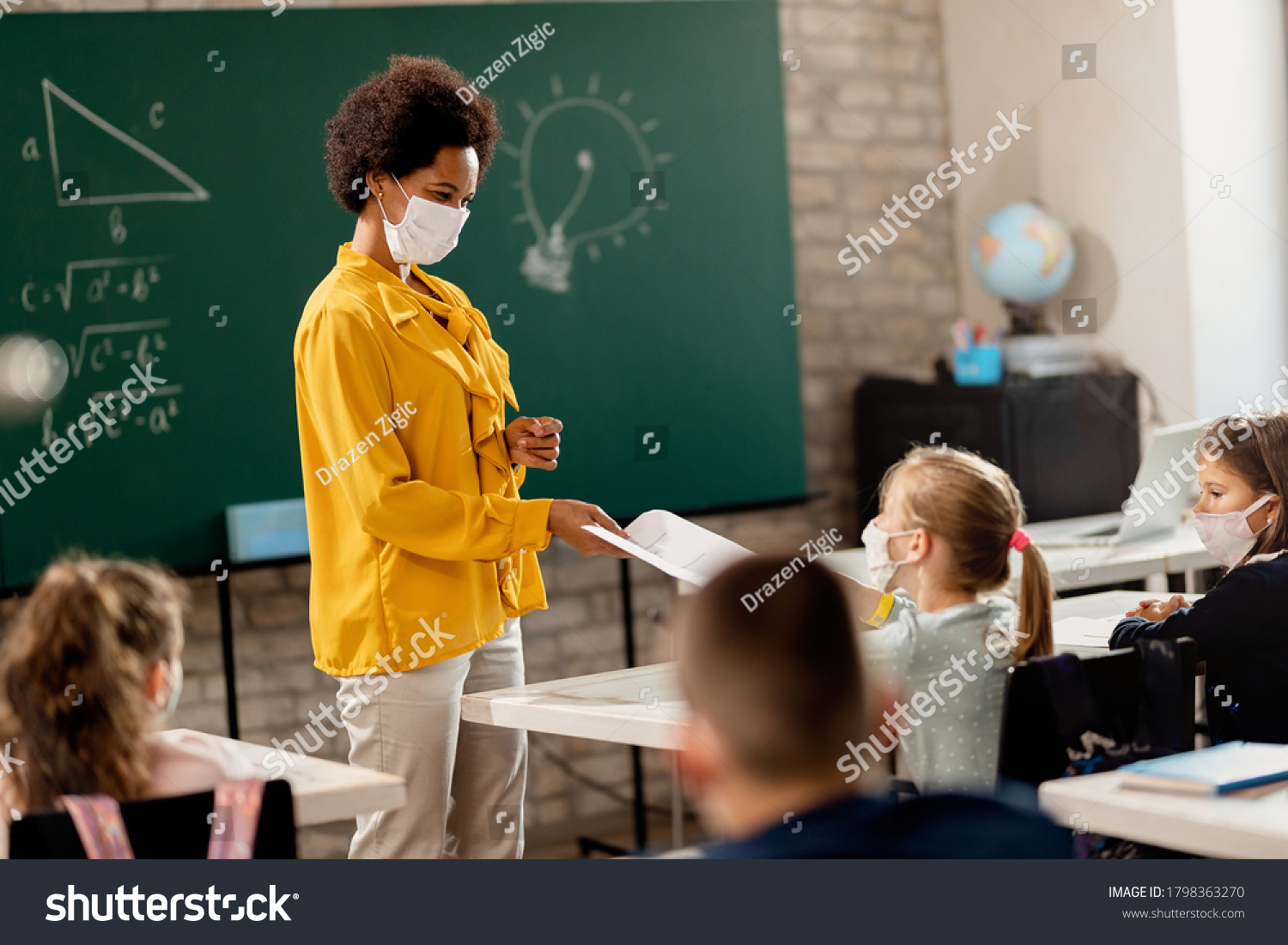 Happy black teacher and her students wearing protective face mask in the classroom. Teacher is giving them their test results.  #1798363270