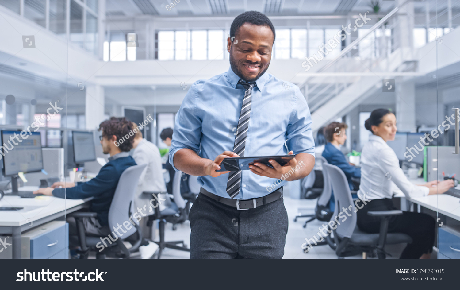 Handsome Young Black Manager in a Shirt Walking Pass His Business Colleagues with a Tablet and Supervise Their Work. Diverse and Motivated Business People Work on Computers in Modern Open Office. #1798792015