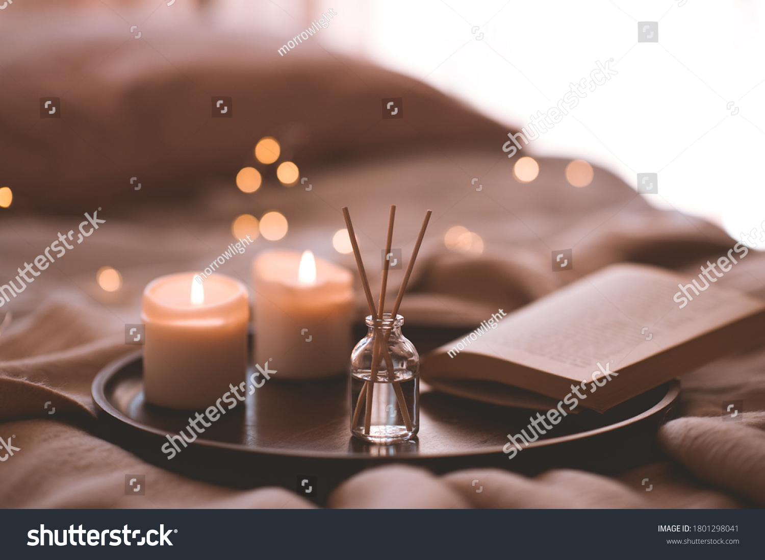 Bamboo sticks in bottle with scented candles and open book on wooden tray in bed closeup. Home aroma.  #1801298041