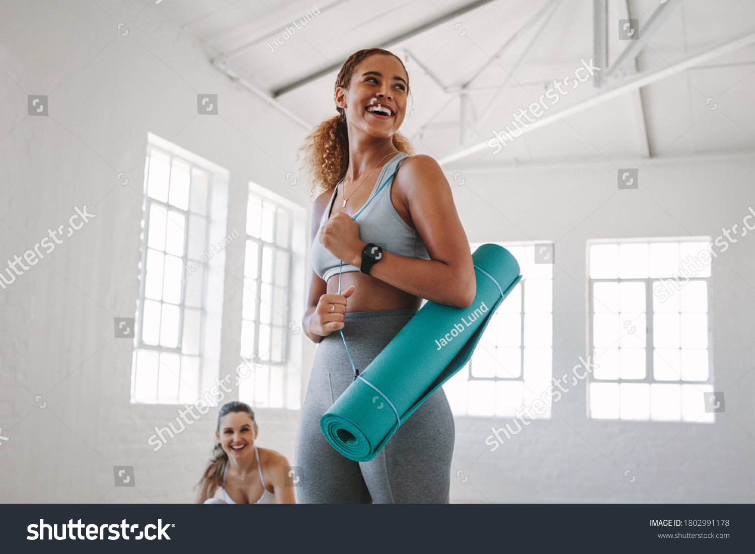 Smiling fitness woman standing in a fitness studio carrying a yoga mat. Portrait of a young woman at a fitness training centre. #1802991178