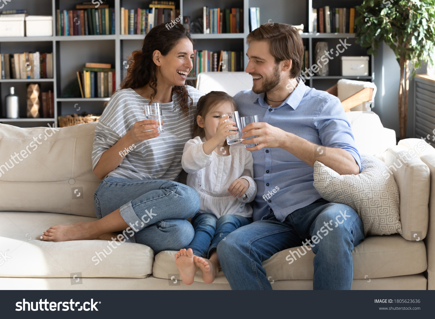 Happy young couple parents teaching little preschool daughter drinking clear water every day. Smiling healthy family holding glasses with pure aqua, enjoying morning daily healthcare habit at home. #1805623636