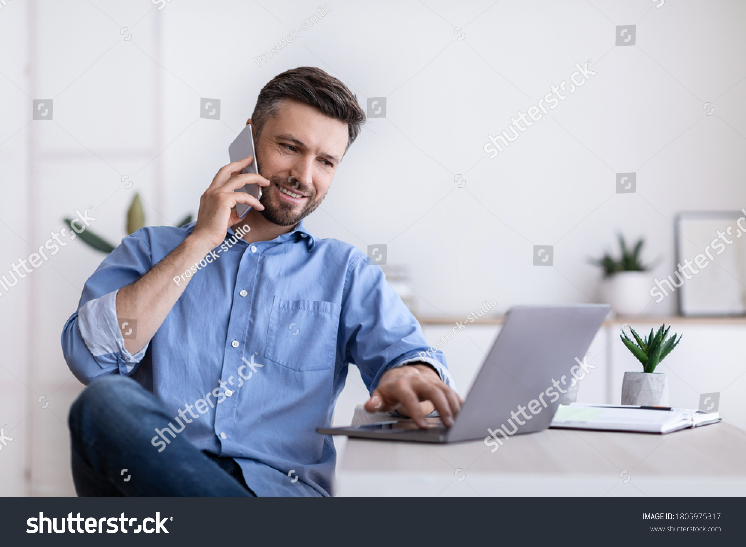 Cheerful Young Man Working Remotely From Home Office With Cellphone And Laptop, Sitting At Desk In Room, Copy Space #1805975317