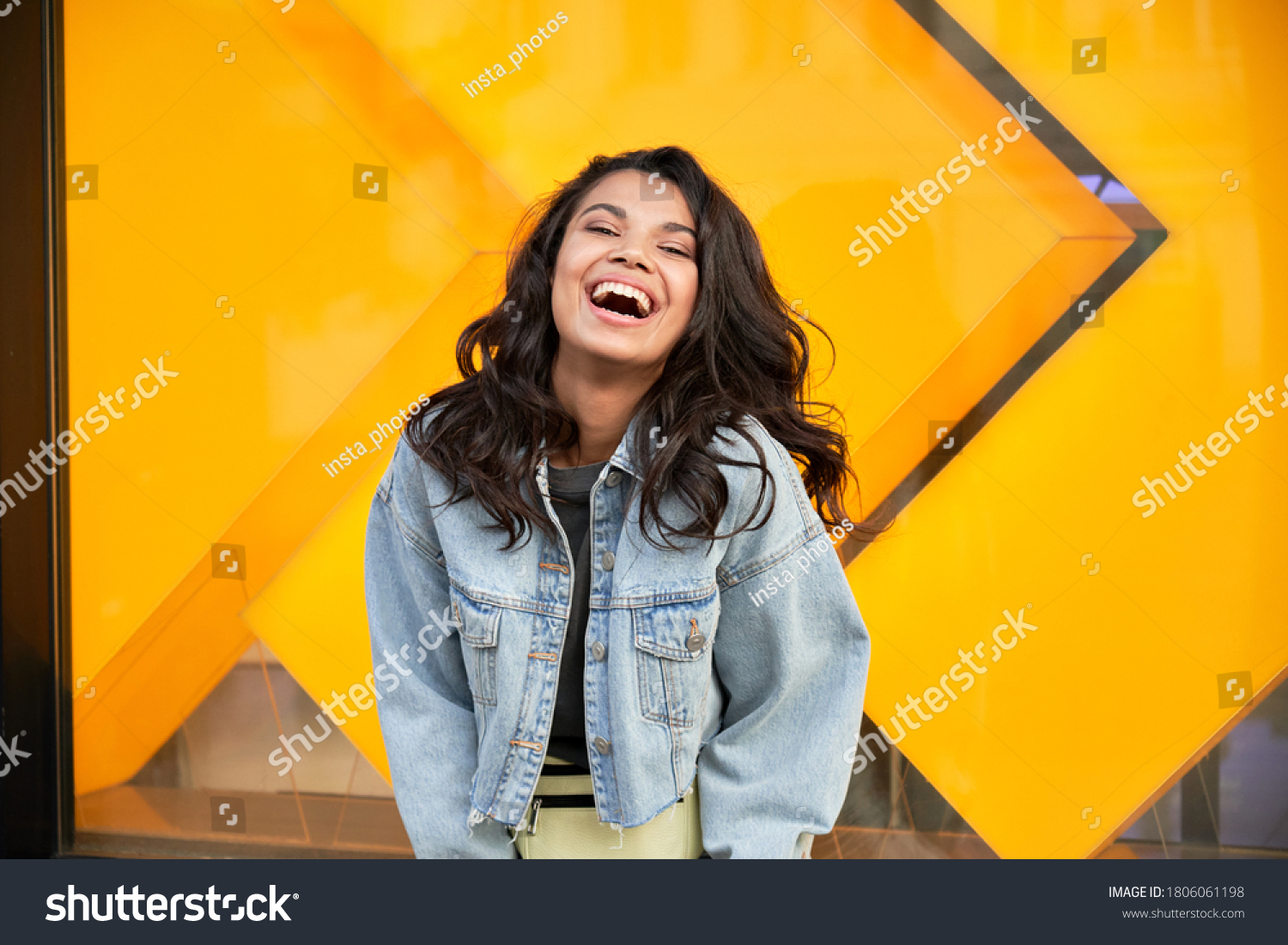 Happy African American woman wearing denim jacket laughing looking at camera standing near city street building. Smiling positive mixed race generation z hipster lady posing outdoor. #1806061198