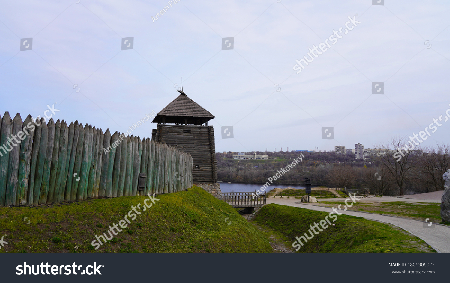 Landscape on a historic temple made of wood in Europe. The fortress is fenced with a wooden palisade against the background of the sky and the river. Thick beams with a sharp top. #1806906022