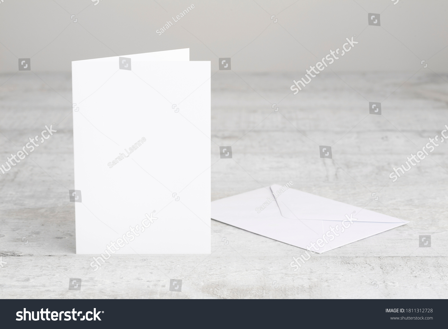 One white greeting card mockup with envelope, standing upright on a white wooden desk. Blank, closed card template.  #1811312728