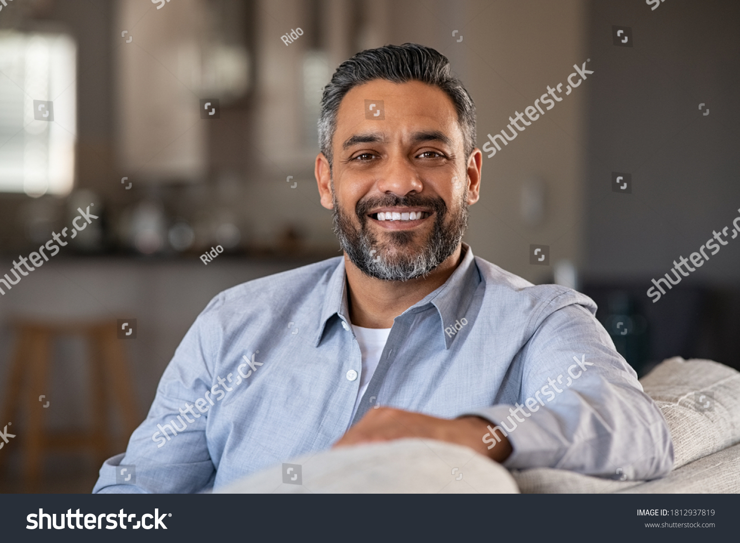 Portrait of happy mid adult man sitting on sofa at home. Handsome latin man in casual relaxing on couch and smiling. Cheerful indian guy looking at camera.  #1812937819