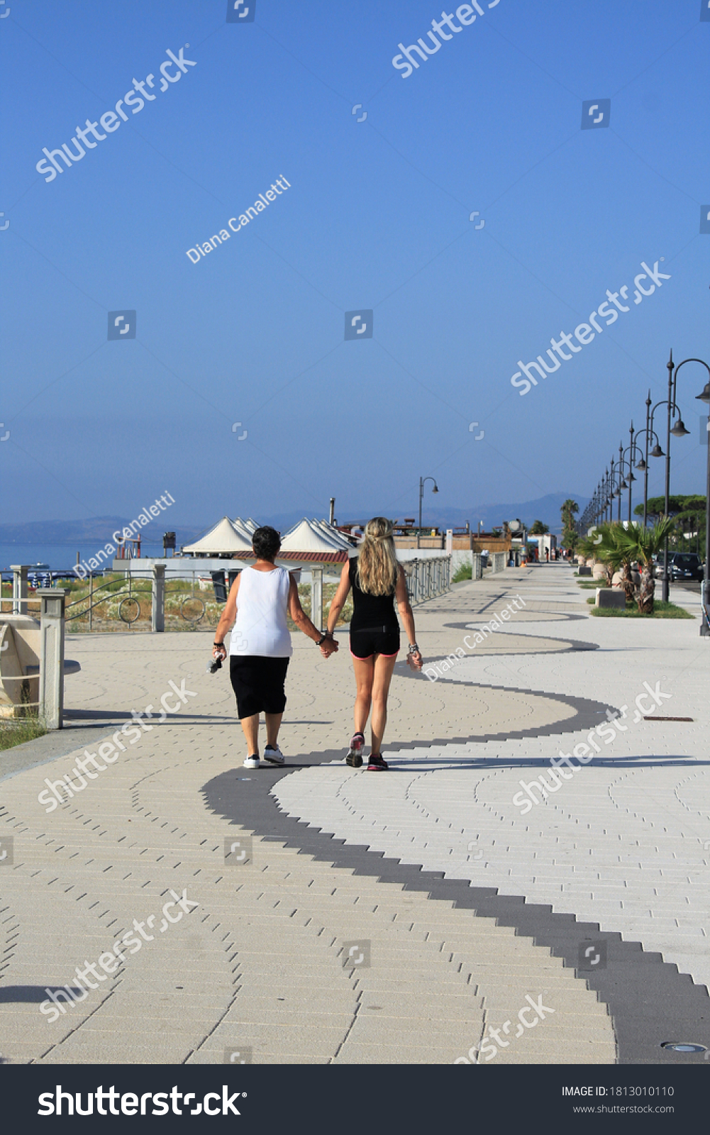 Mother and daughter walk along the promenade of Ardore in Calabria. Walking by holding hands. Two women filmed from behind as they walk. Two people on the seafront. Original corrugated flooring - wave #1813010110