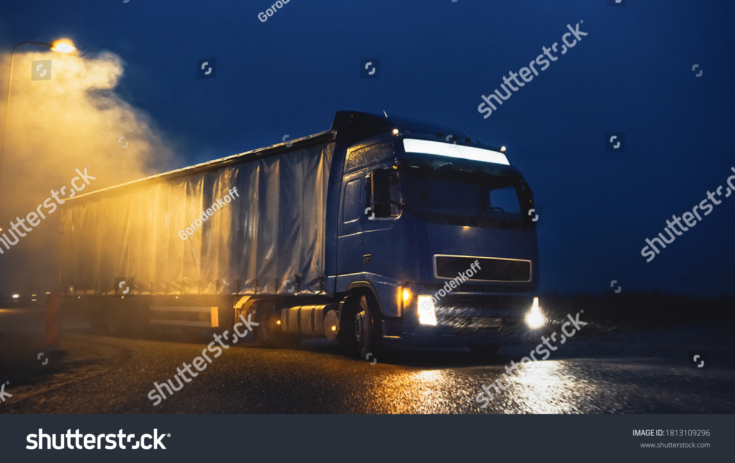 Blue Long Haul Semi-Truck with Cargo Trailer Full of Goods Travels At Night , Turning on Freeway Road, Driving Across Continent Through Rain, Fog Snow. Industrial Warehouses Area. #1813109296