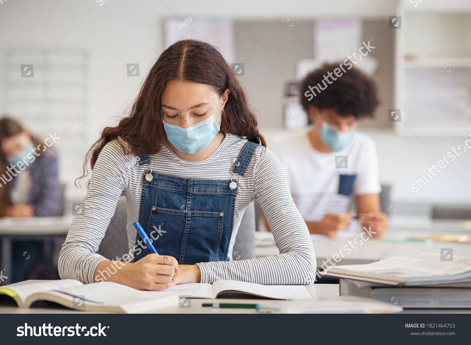 High school student taking notes while wearing face mask due to coronavirus emergency. Young woman sitting in class with their classmates and wearing surgical mask due to Covid-19 pandemic. #1821464753