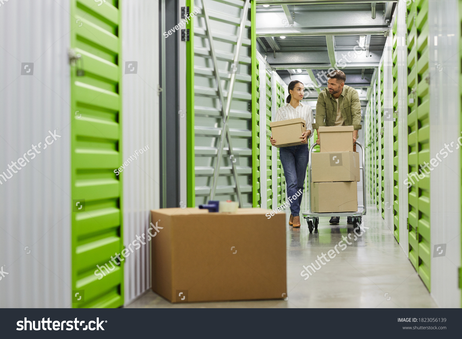 Full length portrait of young couple holding cardboard boxes walking towards camera in self storage unit, copy space #1823056139