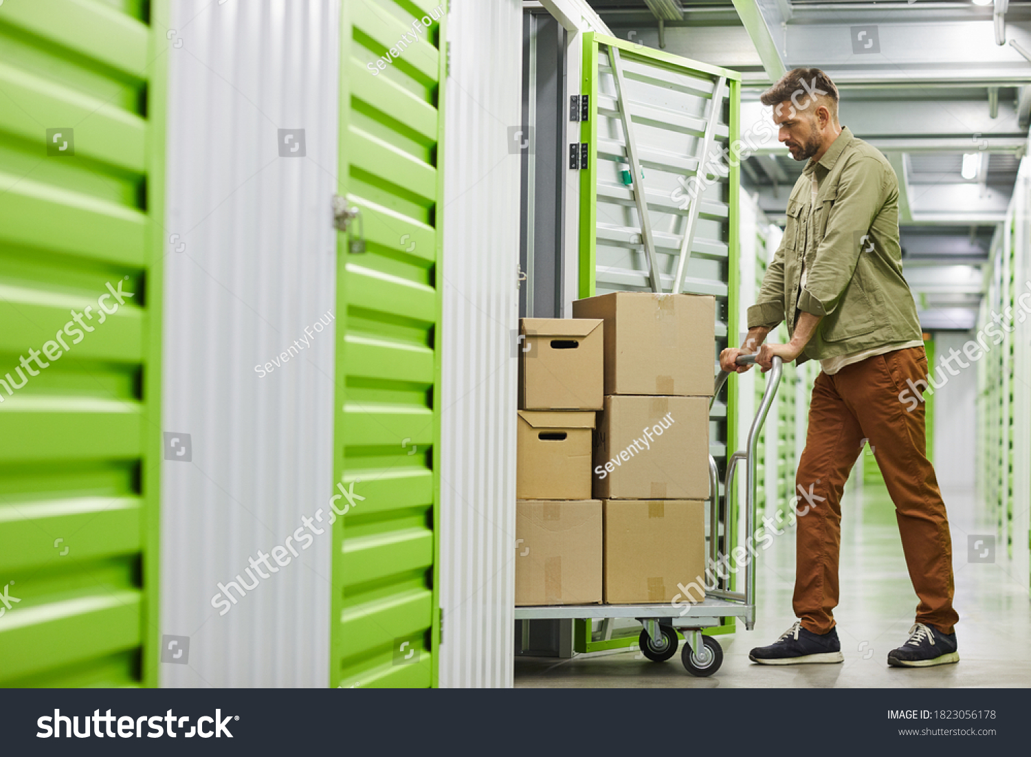 Full length side view at handsome bearded man loading cart with cardboard boxes into self storage unit, copy space #1823056178