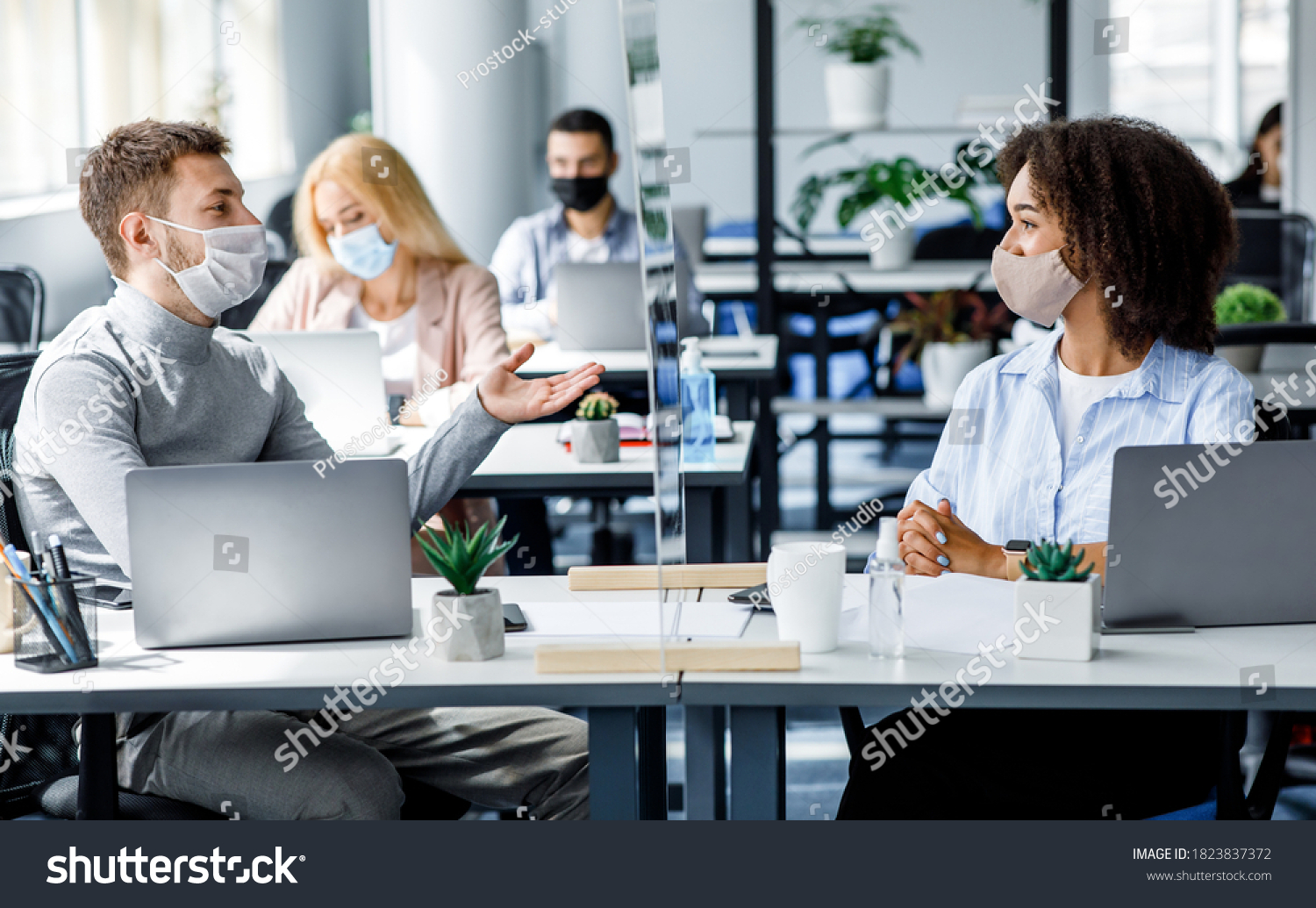 Communication and meeting in office after returning from covid-19 quarantine. Young guy and african american woman in protective mask talking through glass board at workplace with laptops in office #1823837372