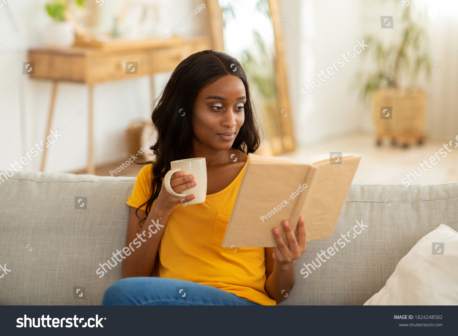 Stay home hobbies concept. Attractive millennial black lady reading book with cup of hot beverage in living room. Focused African American woman with aromatic drink absorbed in intriguing story #1824248582