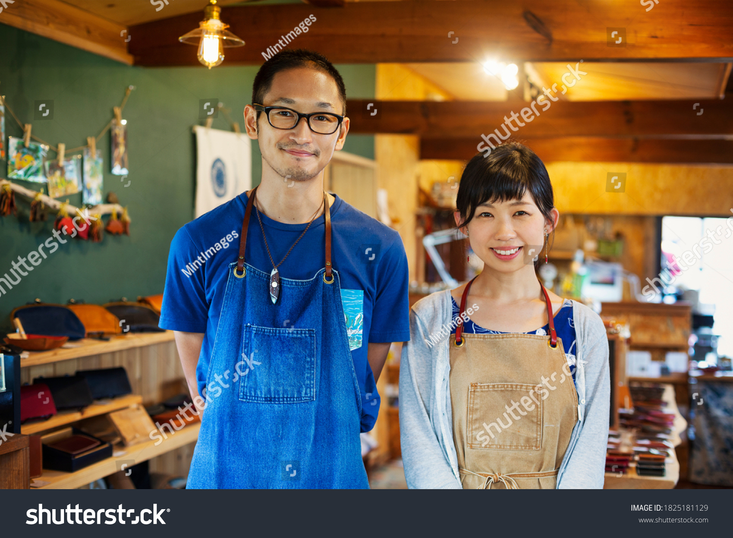 Japanese woman and man wearing blue apron standing in a leather shop, smiling at camera. #1825181129