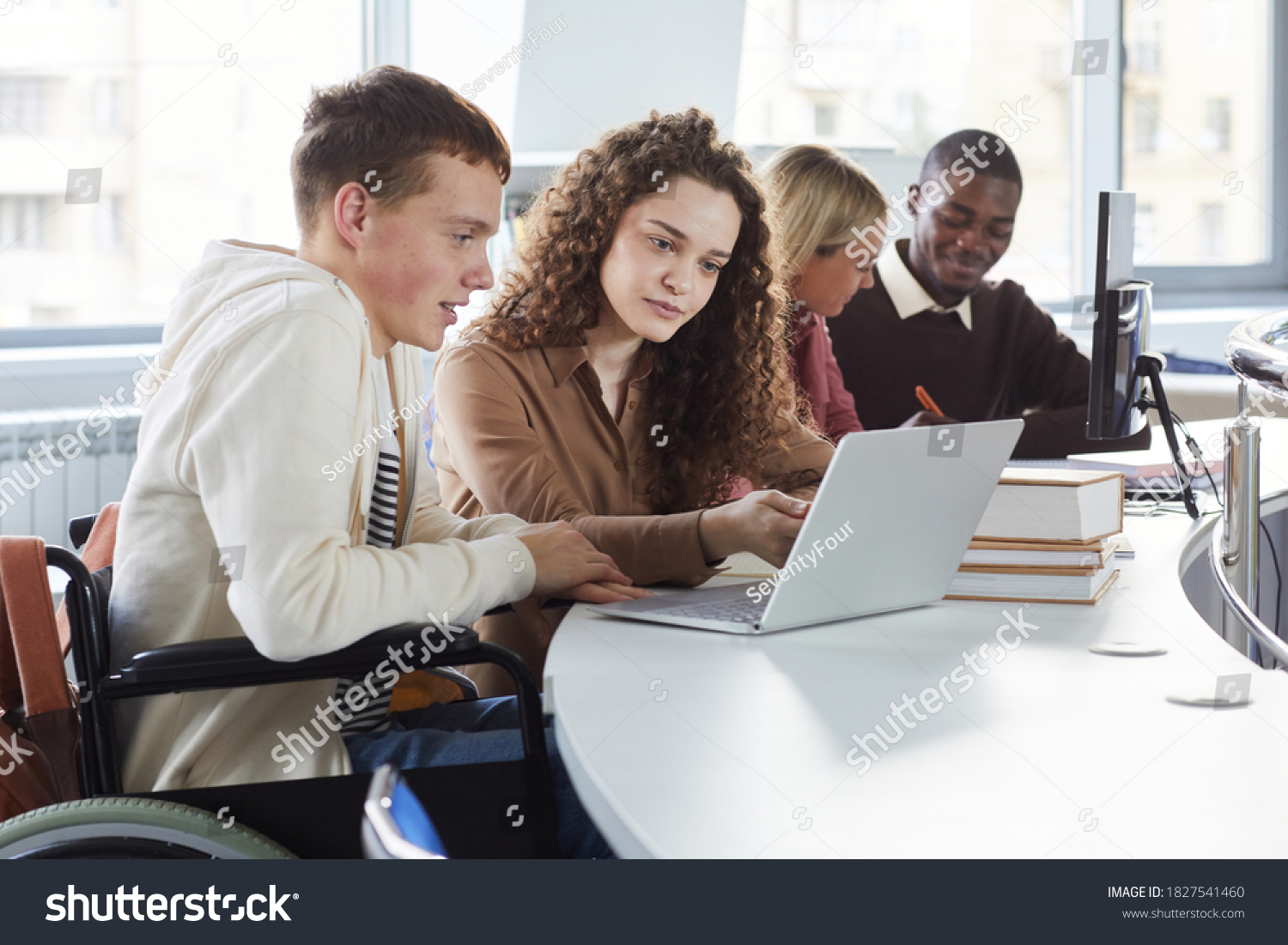 Side view at multi-ethnic group of students using laptop while studying in college, featuring boy using wheelchair #1827541460