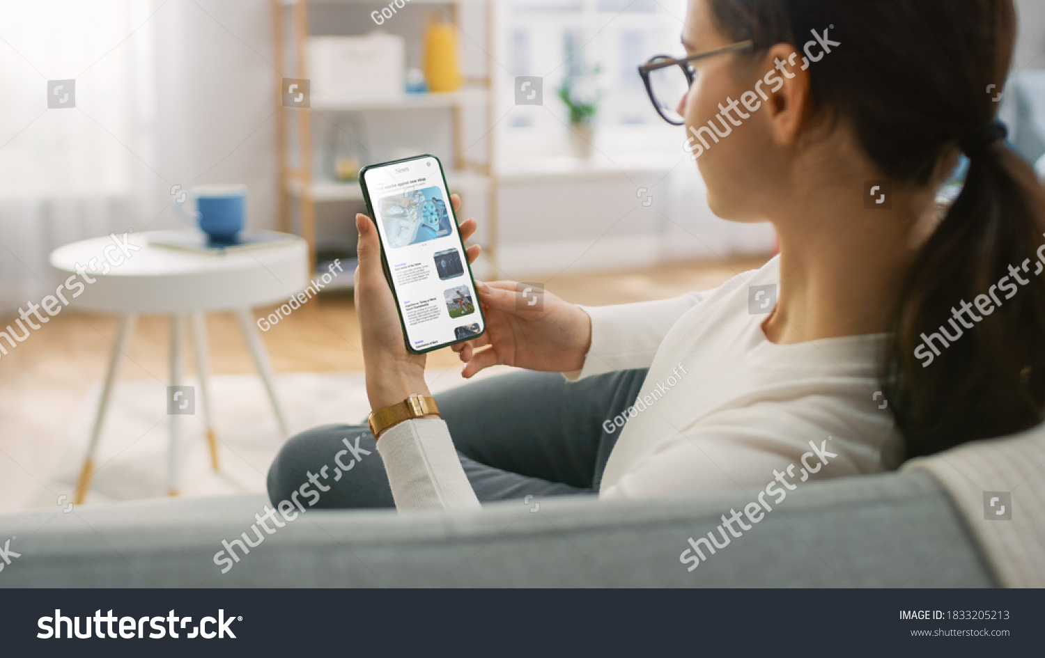 Young Woman at Home Uses Smartphone for Scrolling and Reading News about Technological Breakthroughs. She's Sitting On a Couch in Her Cozy Living Room. Over the Shoulder Shot #1833205213