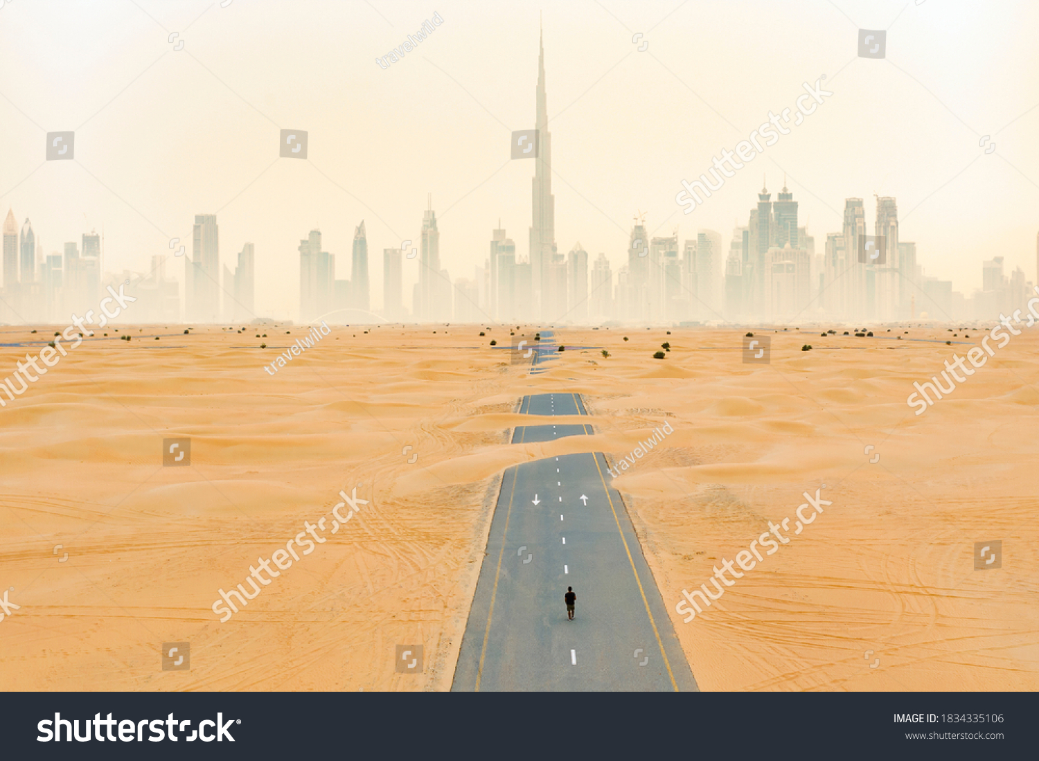 View from above, stunning aerial view of an unidentified person walking on a deserted road covered by sand dunes with the Dubai Skyline in the background. Dubai, United Arab Emirates. #1834335106