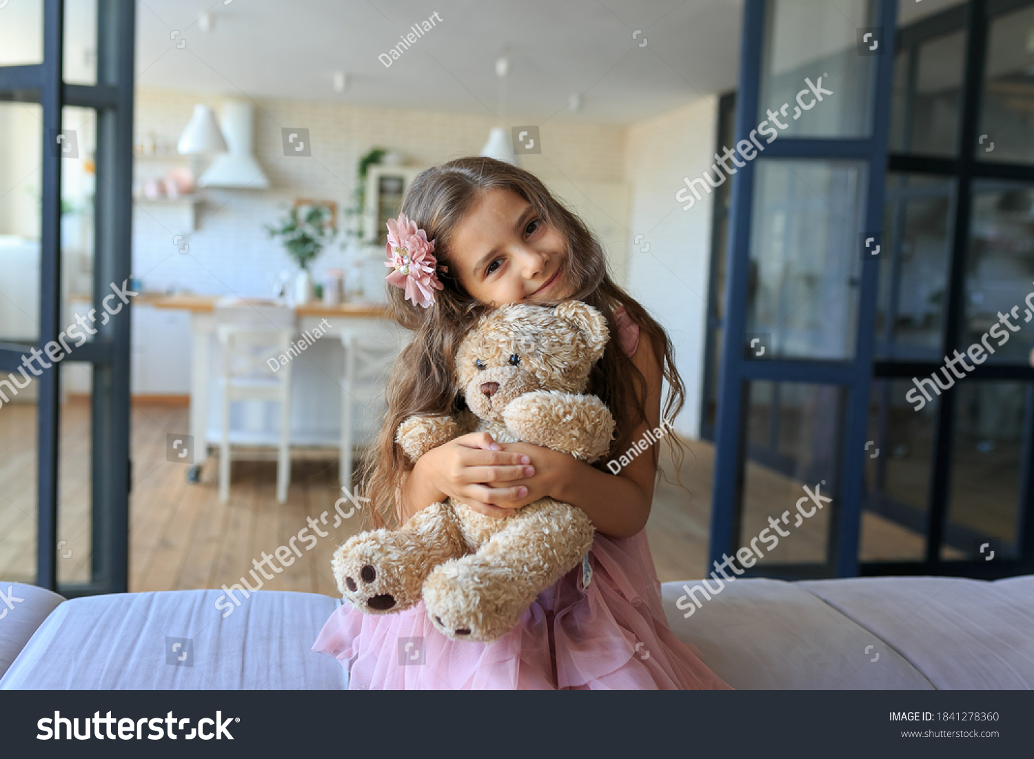 portrait of smiling little girl hugging teddy bear at home. Adorable child with long curly hair with her favorite toy on kitchen background. Happy childhood concept. Cute baby playing with plush bear #1841278360