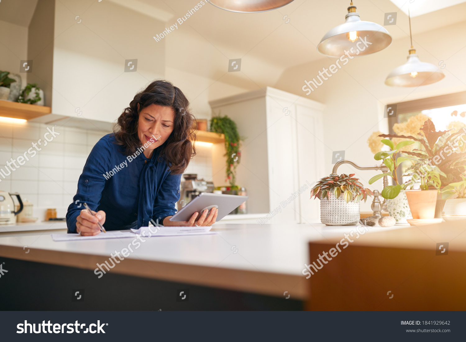Mature Woman With Digital Tablet Reviewing Domestic Finances And Paperwork In Kitchen At Home #1841929642