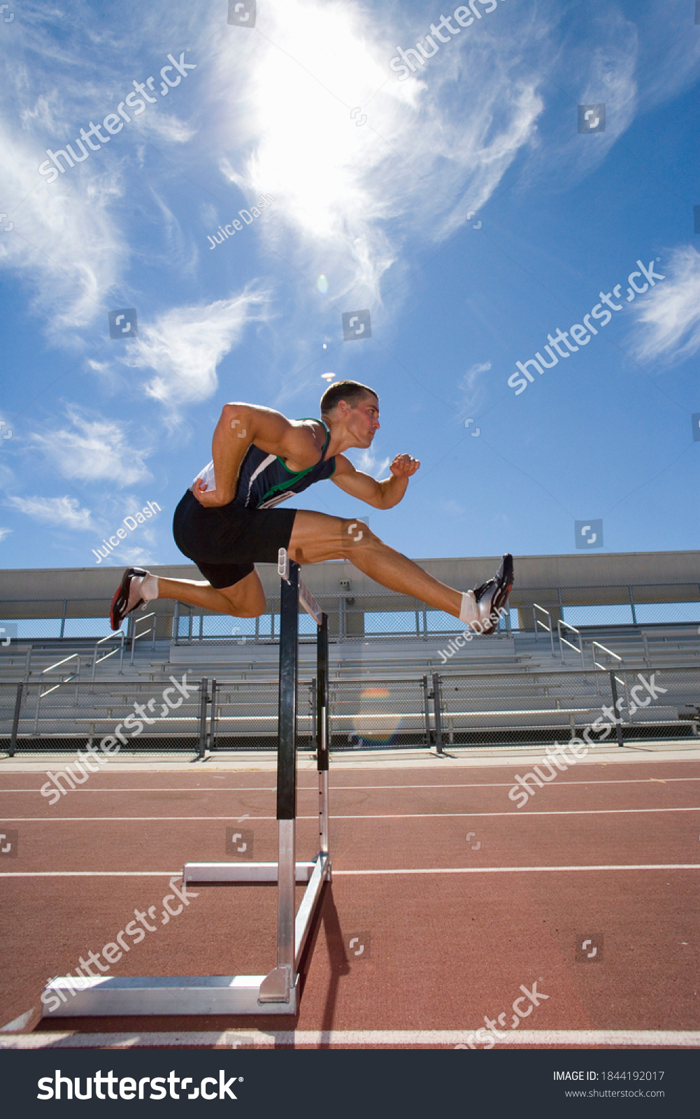 Side view of a male athlete jumping over a hurdle in the race on running track #1844192017