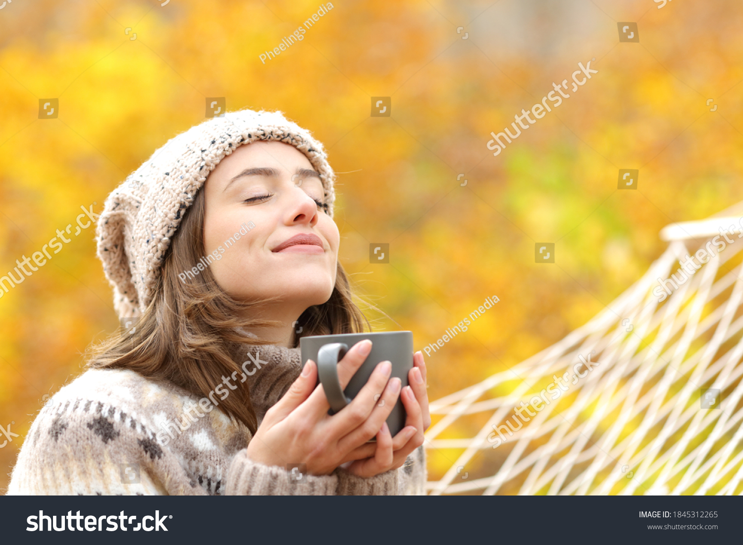 Relaxed woman breaths fresh air holding coffee cup for breakfast sitting on a rope hammock in autumn in a forest #1845312265