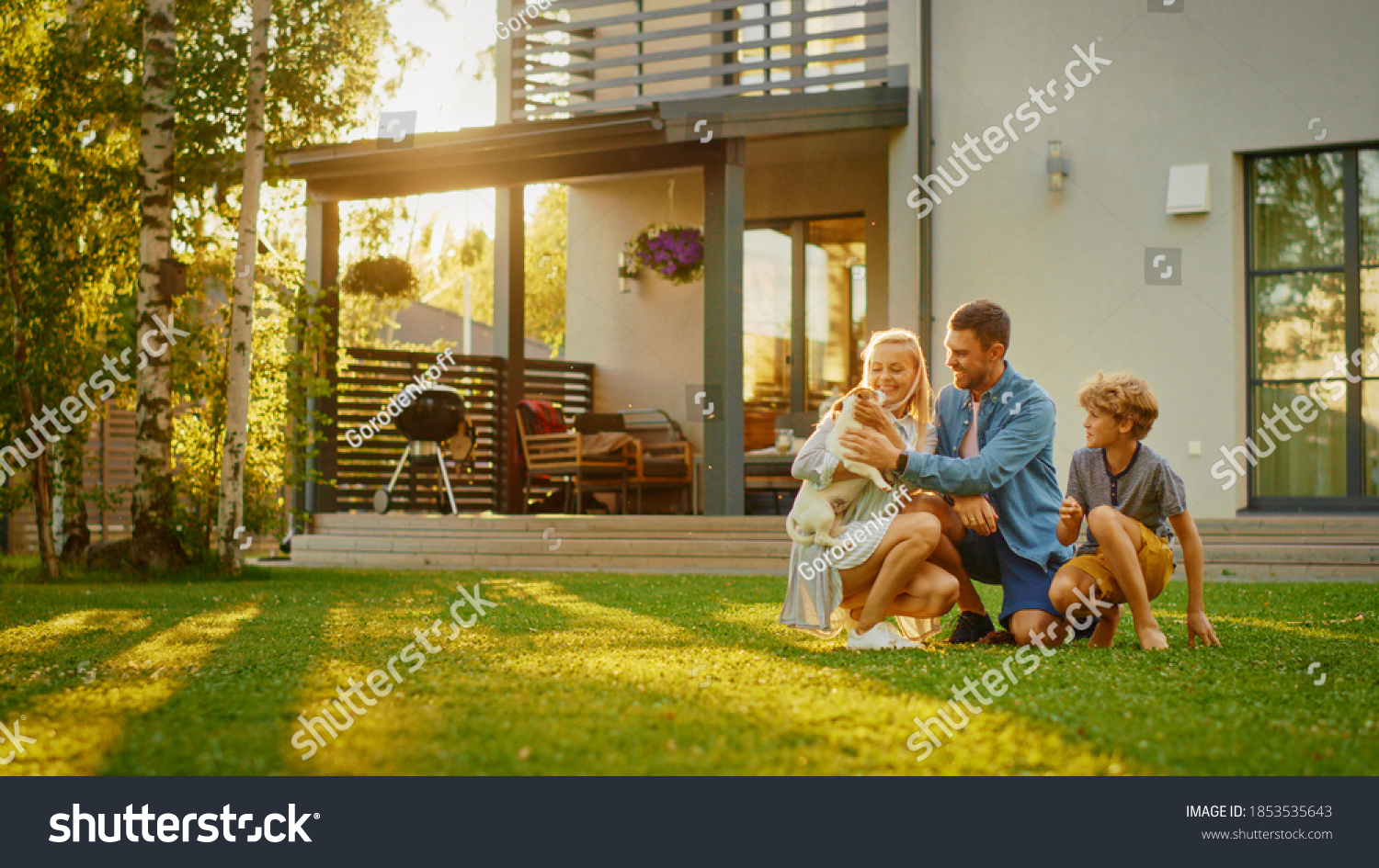 Smiling Father, Mother and Son Pet and Play with Smooth Fox Terrier Retriever Dog. Sun Shines on Idyllic Happy Family with Loyal Pedigree Dog have Fun at the Idyllic Suburban House Backyard #1853535643