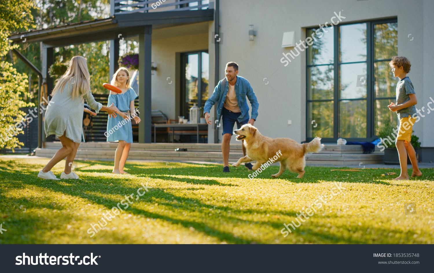 Smiling Beautiful Family of Four Play Fetch flying disc with Happy Golden Retriever Dog on the Backyard Lawn. Idyllic Family Has Fun with Loyal Pedigree Dog Outdoors in Summer House Backyard #1853535748