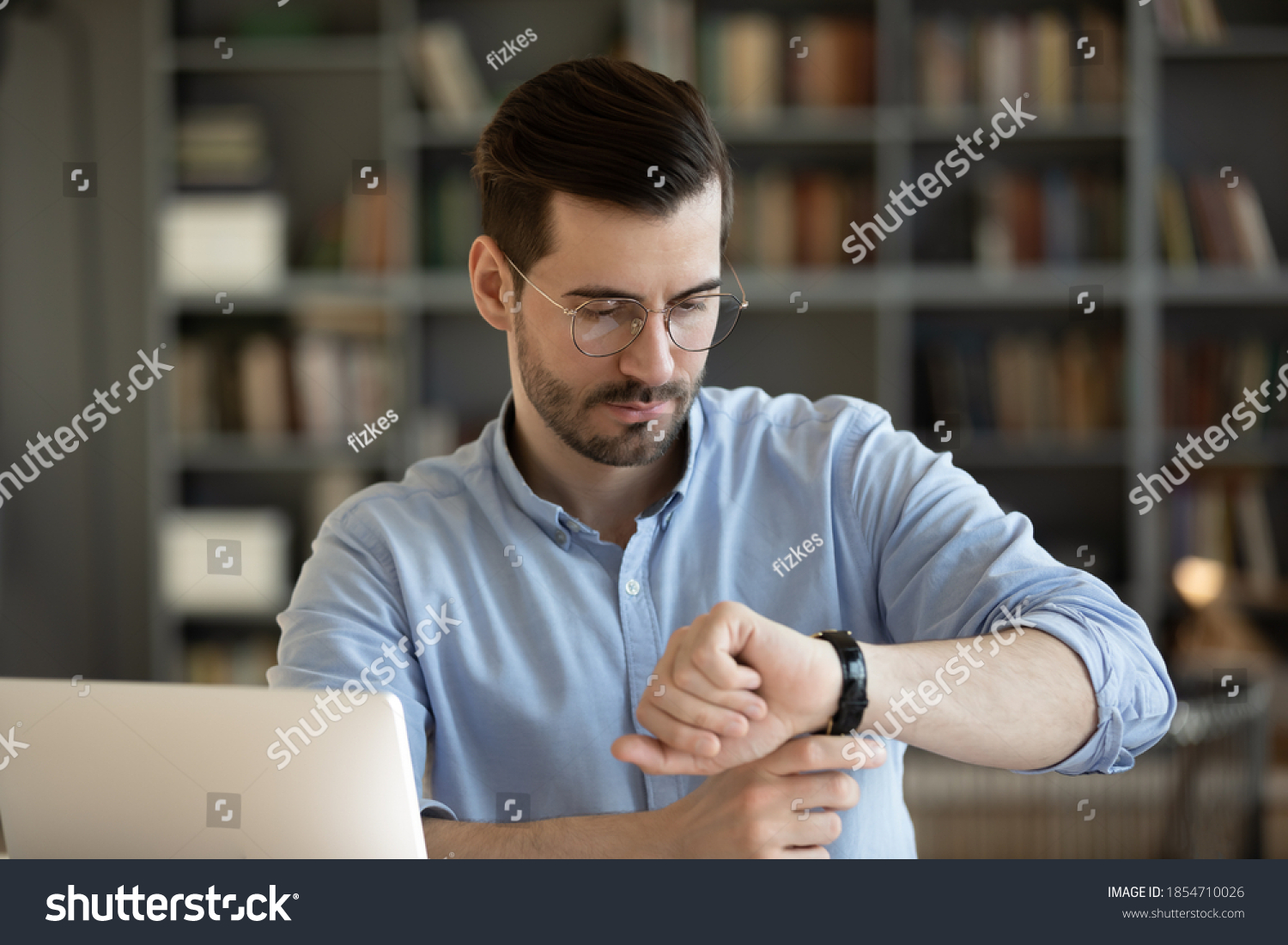 35s businessman wear glasses looks at wristwatch checks time waits for client who is late while sitting at work place desk with laptop. Concept of business, time is money, punctuality and timekeeping #1854710026