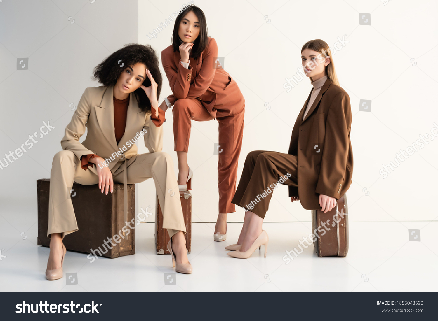 full length of stylish multicultural women in suits sitting on suitcases on white #1855048690