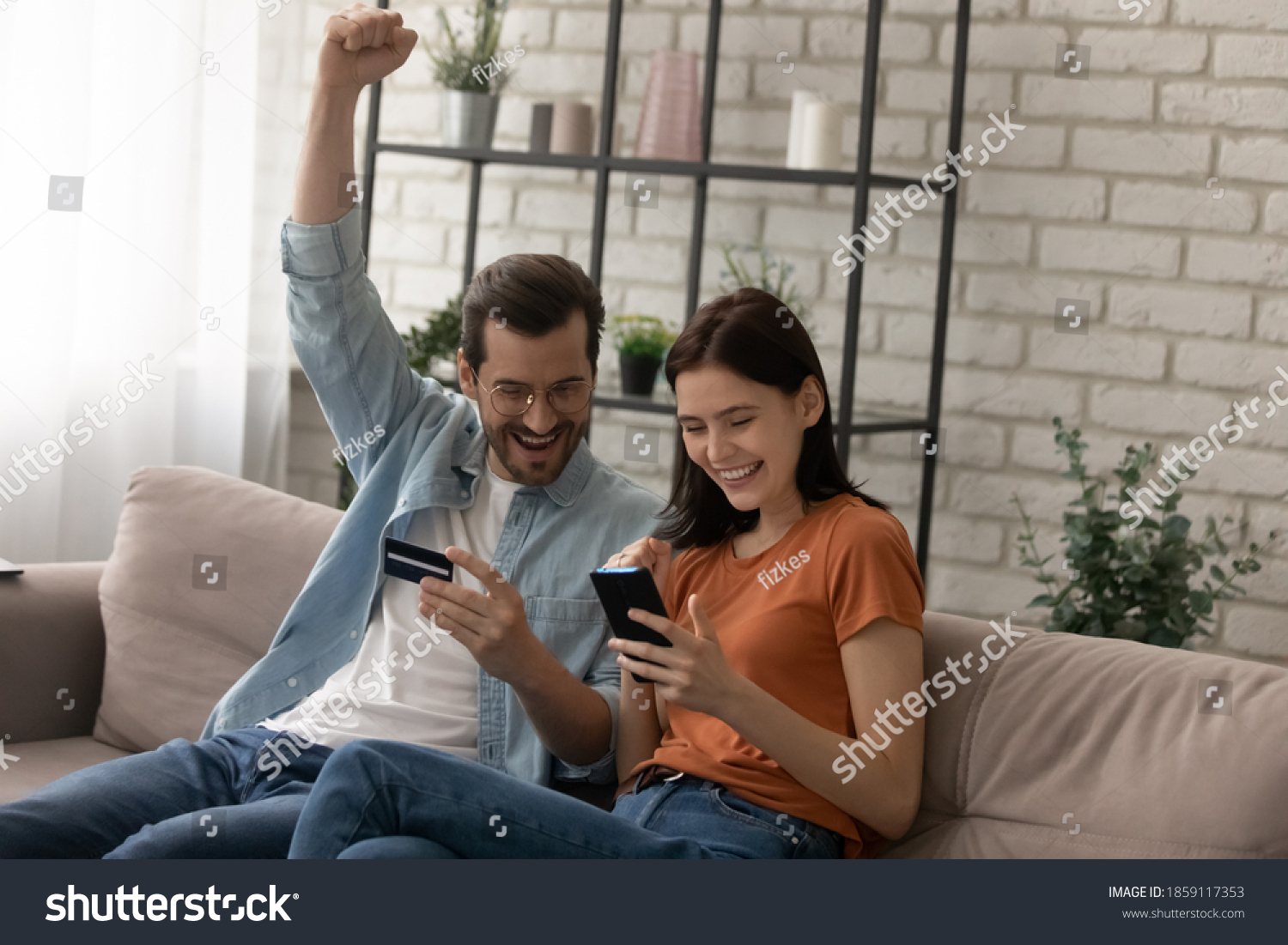 Happy young married family couple involved in online shopping, celebrating getting gift or winning prize in giveaway, paying for goods or services with credit bank card in smartphone application. #1859117353