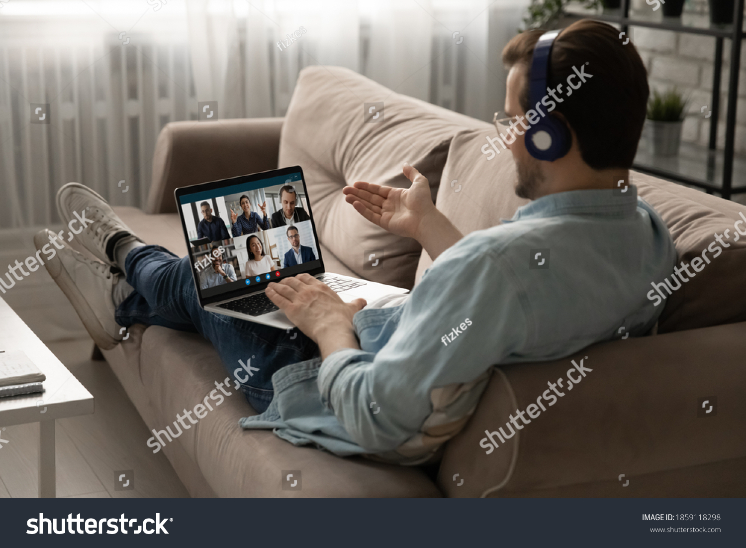 Back rear view young businessman employee in headphones holding video conference online meeting with mixed race colleagues, discussing working issues remotely from home, lying on comfortable sofa. #1859118298