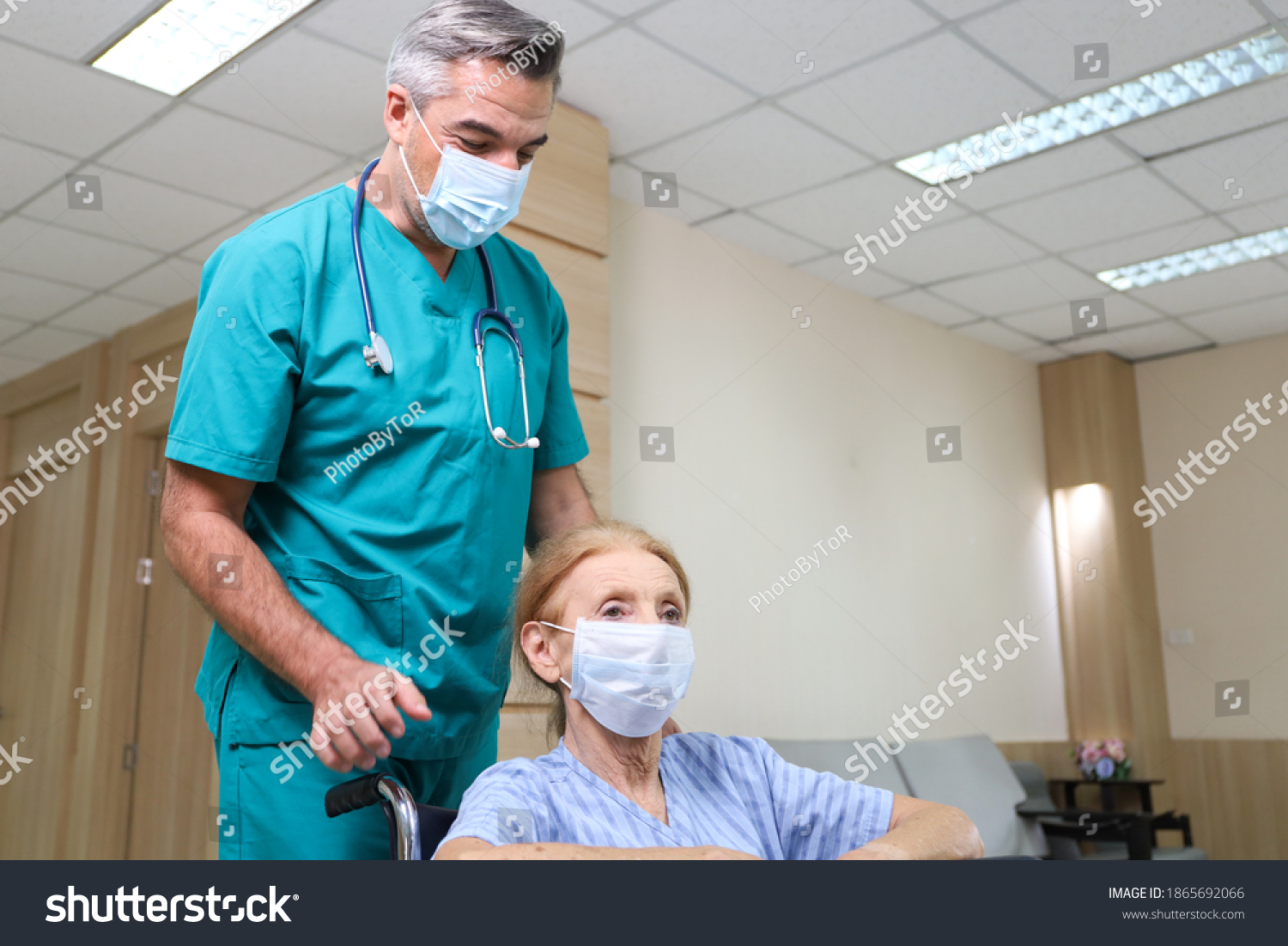 Doctor or assistance staff wear face mask and help a elder patient woman while sitting on wheelchair in the hospital, coronavirus pandemic protection #1865692066