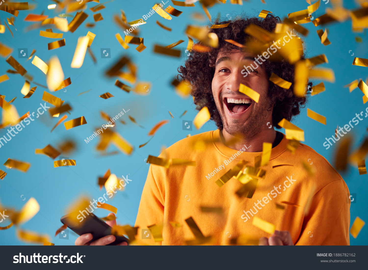 Celebrating Young Man With Mobile Phone Winning Prize And Showered With Gold Confetti In Studio #1886782162